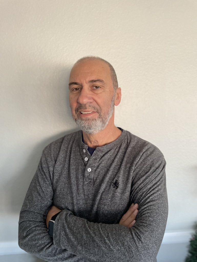 About A man with a gray beard and gray shirt smiling with arms crossed.